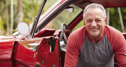 Man sitting in his car, door open, he's seated facing the person taking the camera, legs outside the car. He is smiling